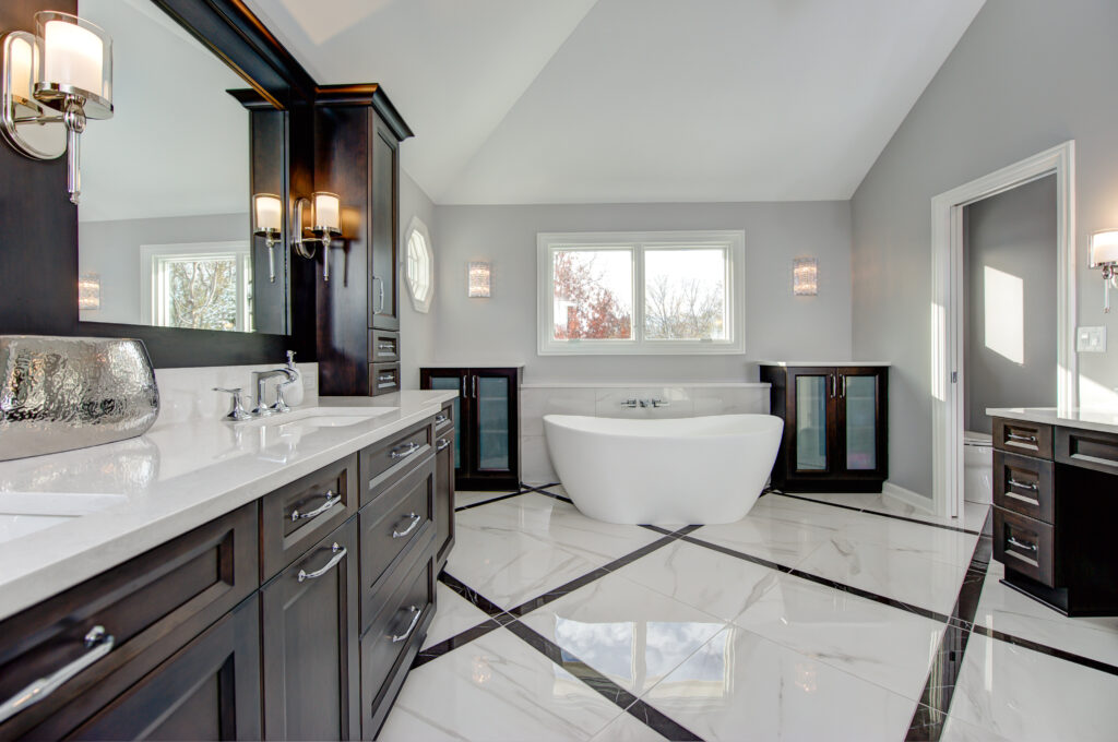 Remodeled primary bathroom with white tile floors, dark cabinets, dual vanity with white countertop, stand-alone white soaking tub at far end of room.