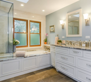 Coastal style bathroom remodel with soaking tub, white cabinets, gray stone countertop, pale blue walls, privacy windows over tub.