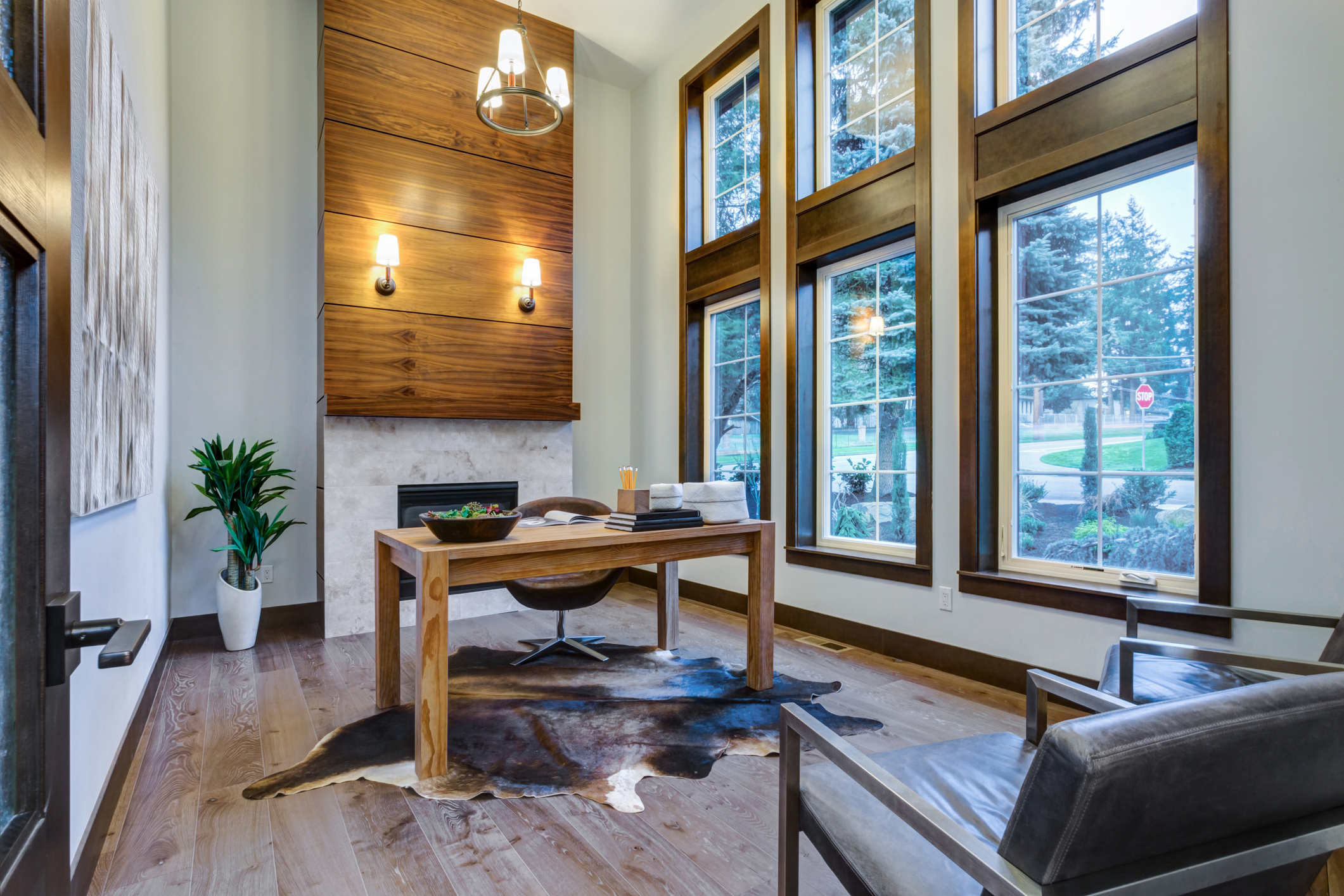 Home office with light wood floors, three large wood-framed floor-to-ceiling windows with grids along right wall, fireplace along far wall, light wood desk in the middle, on a bear skin rug. Vaulted two-story ceiling height.