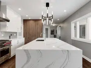 Modern kitchen remodel with large island with white marble waterfall countertop. White cabinets along left wall, light gray wall on right, modern pendant light above island.