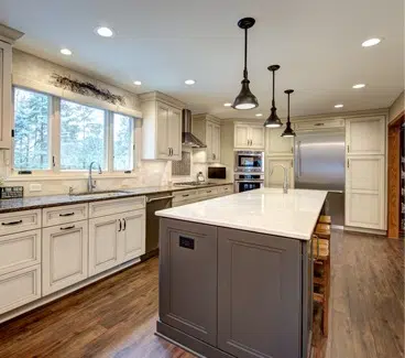 Remodeled large kitchen with wood floors, large island with brown cabinets and white stone countertop. White cabinets along left wall with row of three windows over sink. Recessed and pendant lighting.