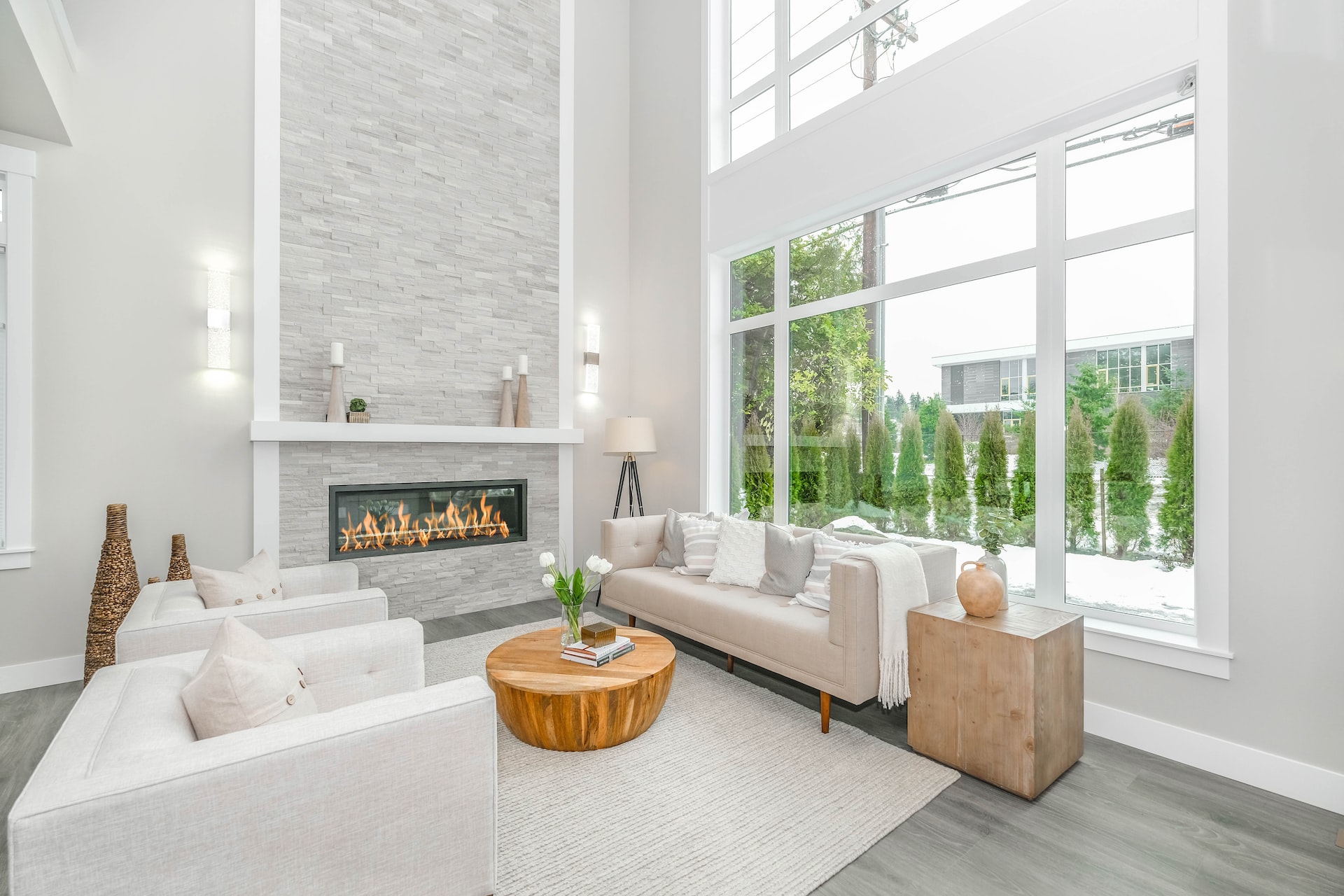 Modern electric fireplace with white brick/tile facade at end of modern living room with gray wood floors, white couches and rug, and two-story wall of windows to the right.