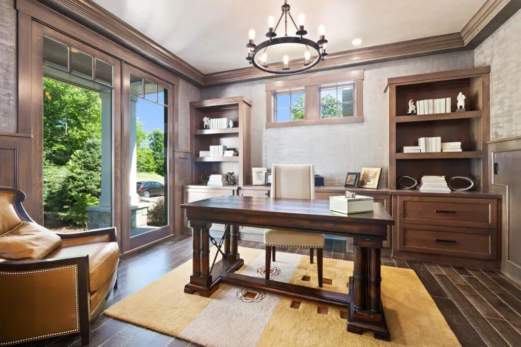 Home office remodel with dark wood floors, large floor-to-ceiling windows on left wall, built-in shelving and drawers on far wall, and large wood desk in center of room.